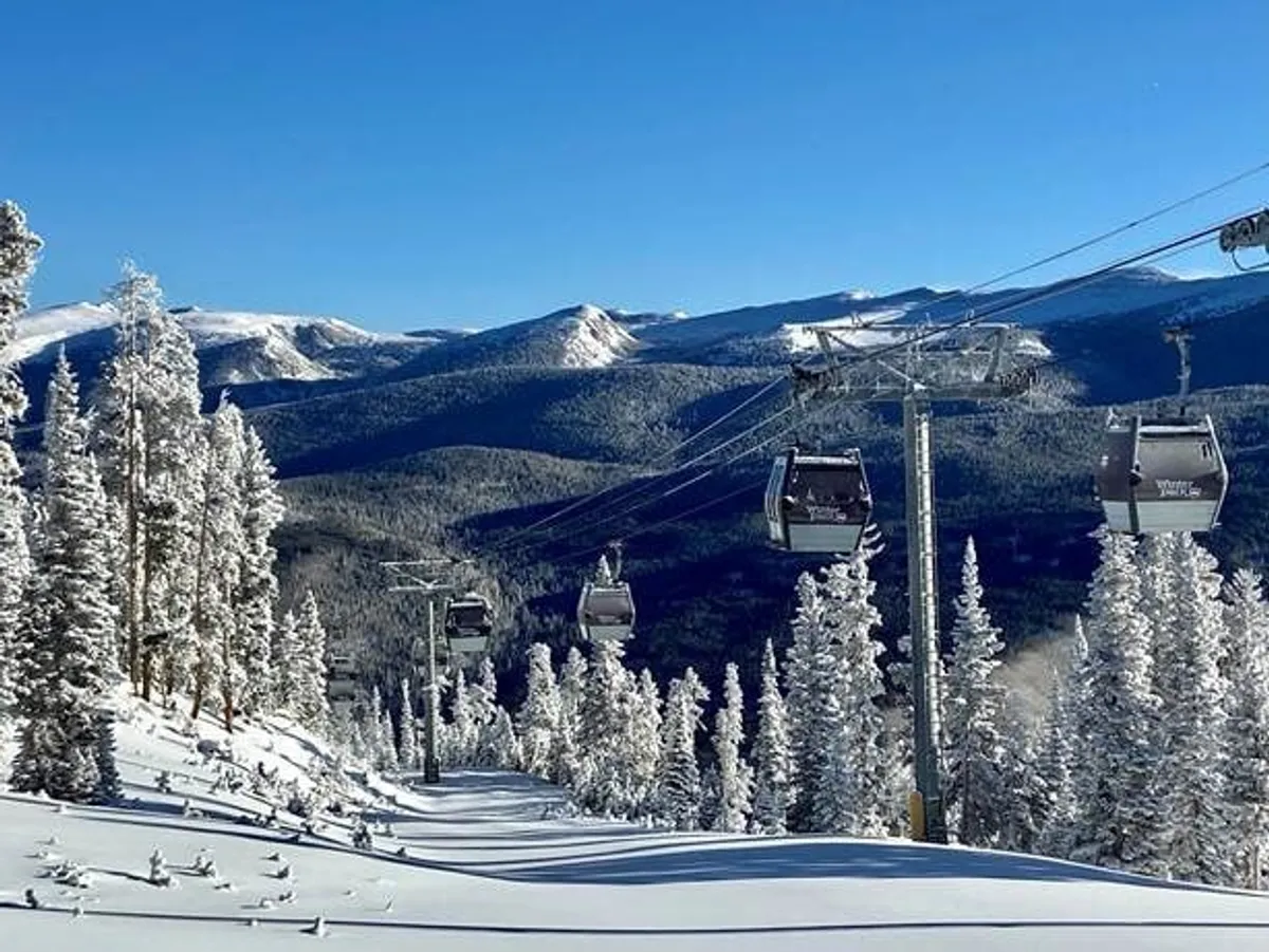 A sweeping view of snow-covered Rocky Mountains under a clear sky, with ski lift gondolas gliding above pine trees. The landscape captures the serene beauty and adventure opportunities that define the Winter Park area.