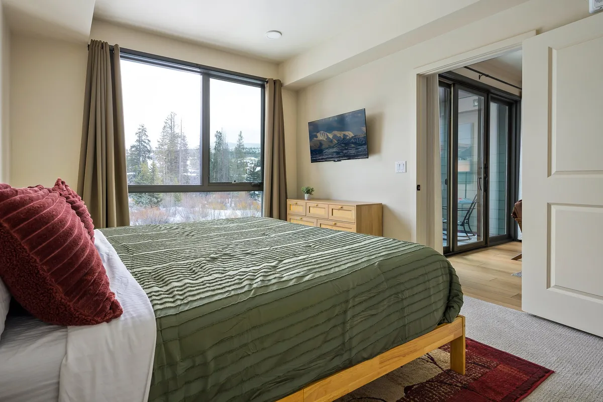 A cozy bedroom featuring a bed with a green coverlet, a large window dressed with curtains that lets in ample natural light, a wooden dresser, and a wall-mounted TV. The soft lighting and thoughtful décor create a tranquil and inviting atmosphere ideal for relaxation.