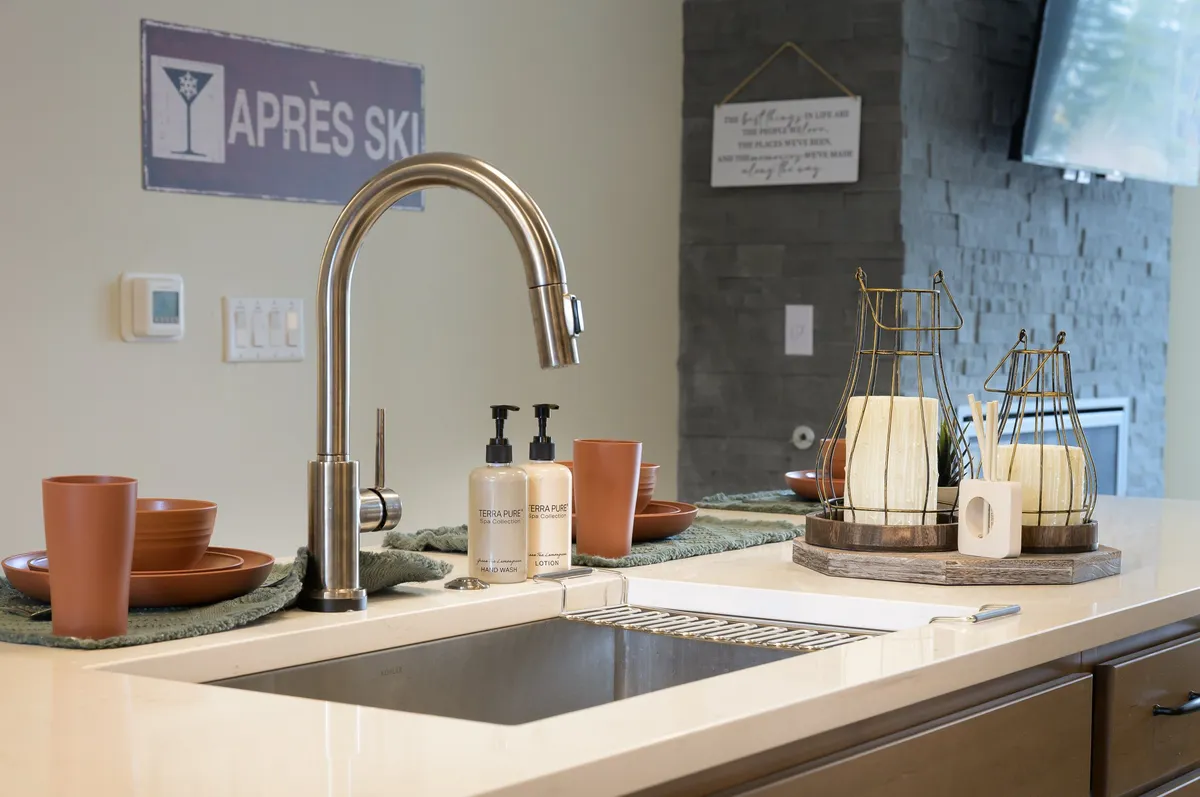A stylish kitchen scene featuring a modern faucet, a prominent 'après ski' sign, a wooden serving tray with candles, and ceramic bowls. The warm lighting and cozy setup suggest a welcoming space to gather after outdoor adventures.