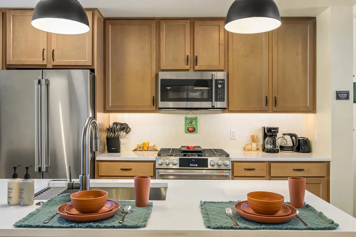 This image captures a contemporary kitchen featuring a spacious island with table settings, stainless steel appliances, and warm wooden cabinets. The setup radiates a welcoming, functional atmosphere perfect for family cooking or casual meals, showcasing the modern appliances and ample prep space highlighted in the section.