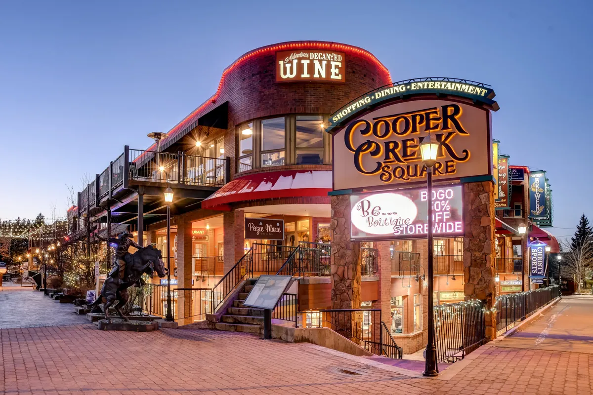 A festive, bustling outdoor area featuring the Cooper Creek Square sign, decorative wine signage, outdoor seating, and a horse sculpture. The space is lively with hints of dining, shopping, and community events.
