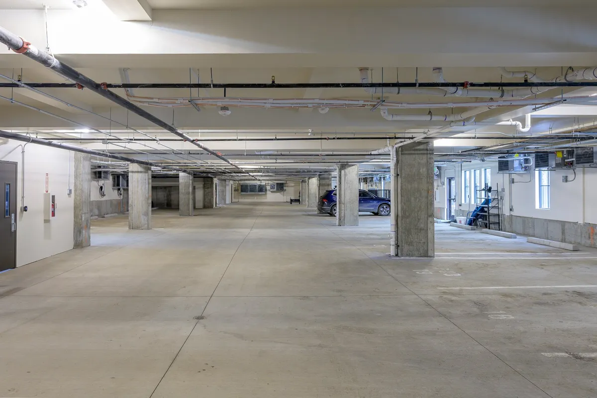 An interior view of an assigned garage with concrete pillars, lighting fixtures, and several cars parked in marked spots. The clean, well-lit space emphasizes convenience and security for residents arriving home or preparing for outings.