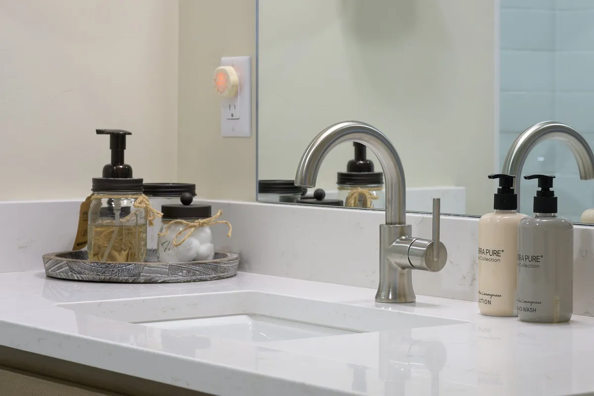 A serene bathroom vanity area featuring a faucet, soap dispenser, and a decorative tray. The mood is calm and inviting, with an emphasis on personal care and relaxation. The image hints at the use of organic, hotel-quality soaps and environmentally-conscious amenities.
