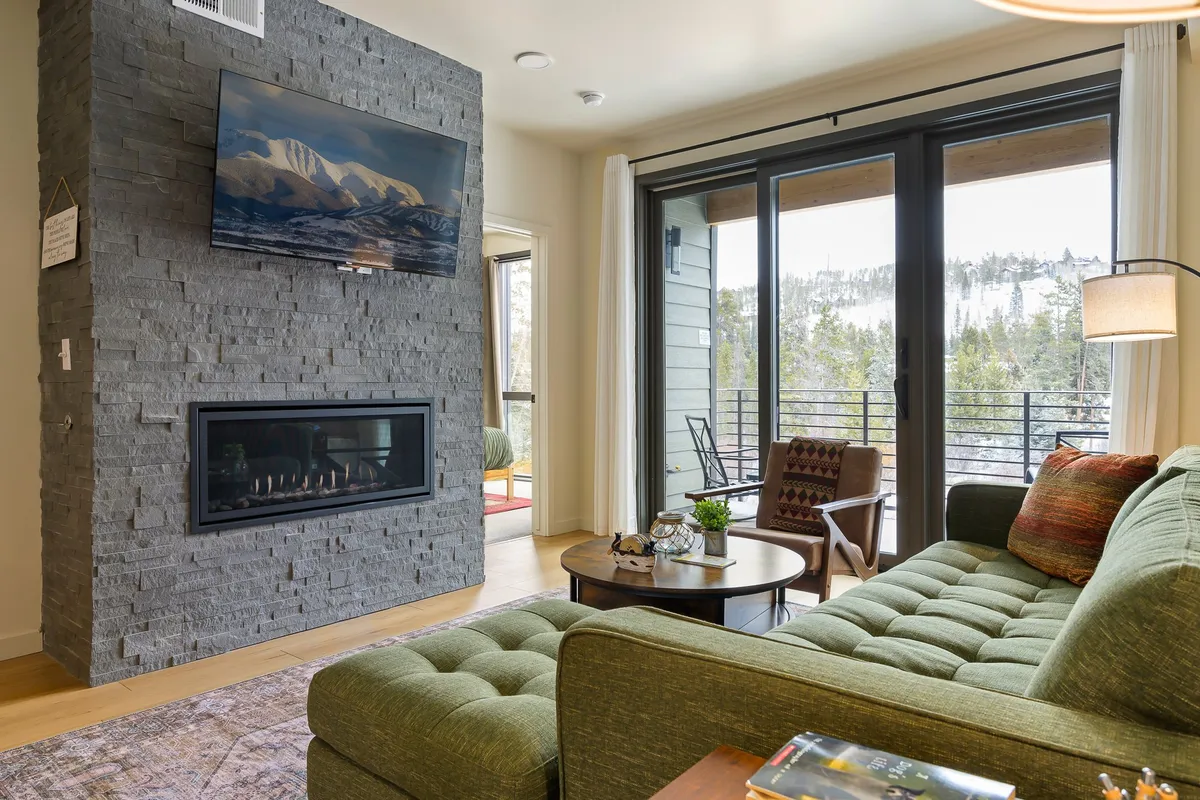 A cozy living room scene featuring a stone fireplace and large windows that offer sweeping mountain views. The room is anchored by a green sofa and a wooden coffee table, with natural light pouring in, creating a serene atmosphere. The space feels closely connected to the outdoors, inviting relaxation after a day spent exploring nature.