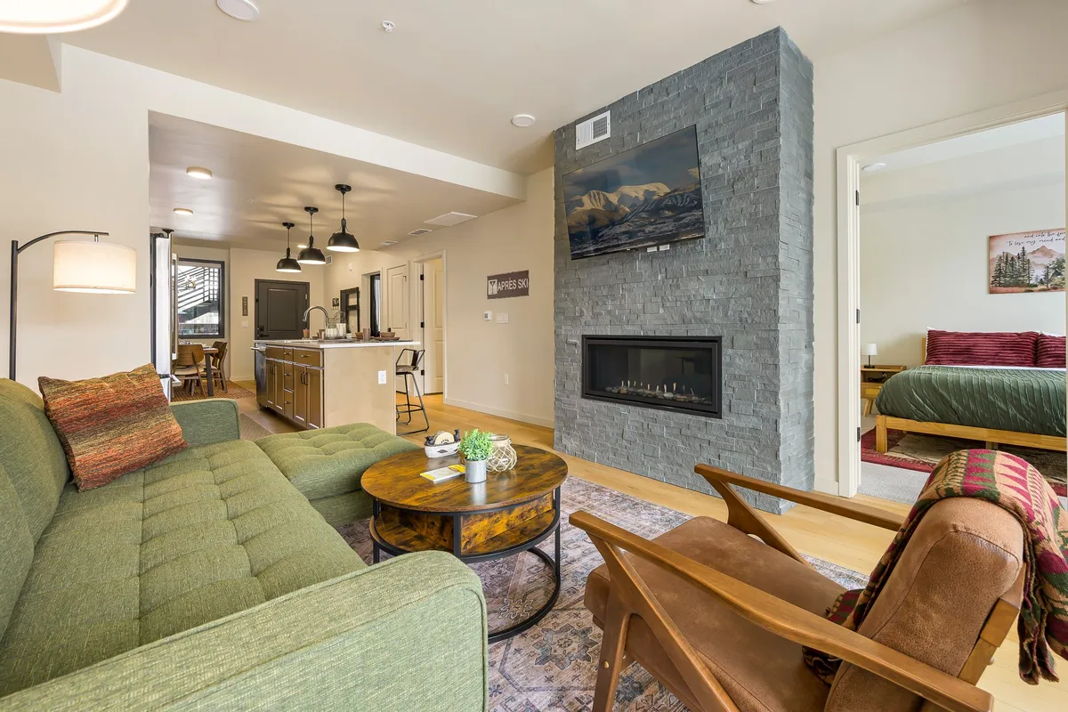 A spacious living area featuring a plush green sofa, stone fireplace, wooden coffee table, a flat-screen TV, and a dining area extending into the background. Large windows allow natural light to pour into the room, accentuating the cozy, inviting atmosphere ideal for gatherings and relaxation.