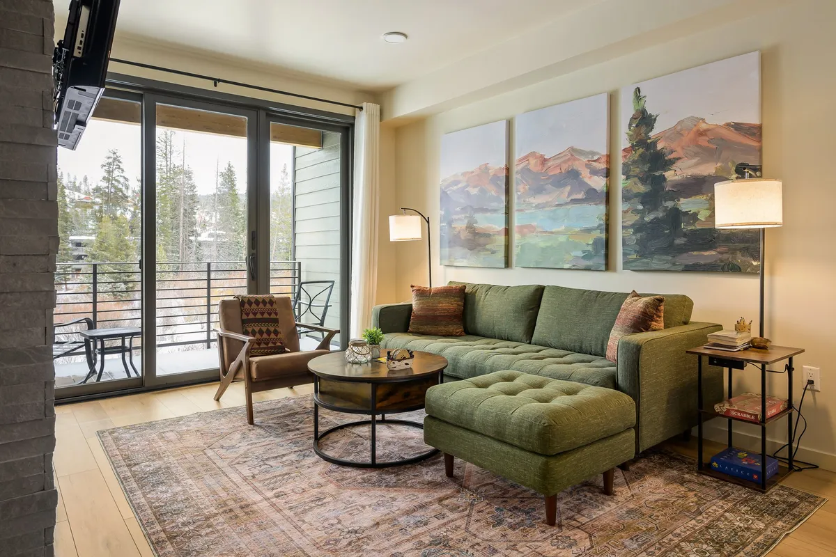 A spacious, comfortable living area with a green sectional sofa, a coffee table, artwork on the wall, and large windows letting in natural light. The setting is perfect for relaxing, entertaining, or enjoying the changing seasonal views outside.