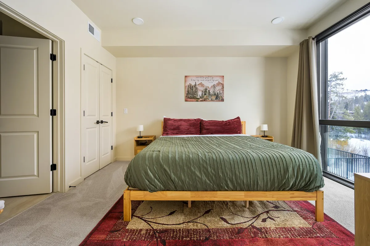 A calm and serene bedroom with a bed dressed in green bedding and red accent pillows. A large window invites in natural light and offers scenic views, complemented by a painting on the wall. The layout encourages leisurely mornings and restful nights.