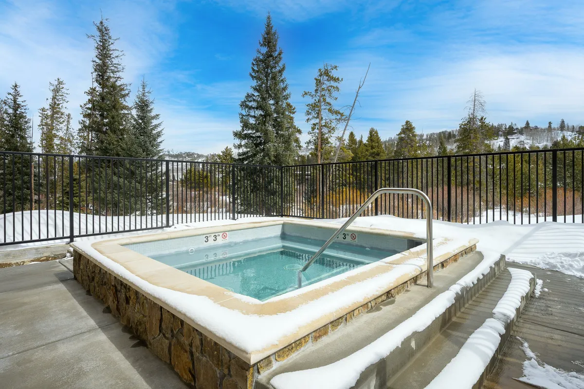 An inviting outdoor hot tub surrounded by snow with distant trees in the background, creating a serene yet playful winter landscape. The setting encourages relaxation and socializing after days spent exploring or playing, and hints at seasonal activities like sledding or snowball fights.
