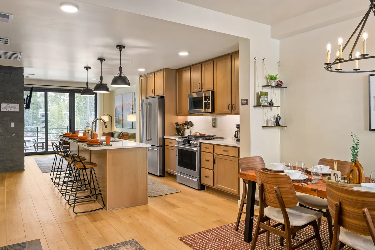 A bright, open-concept kitchen and dining area with a central kitchen island, modern dining table, and expansive windows overlooking the outdoors. The space is filled with natural light and set up for cooking and hosting family meals, capturing the communal heart of the home.