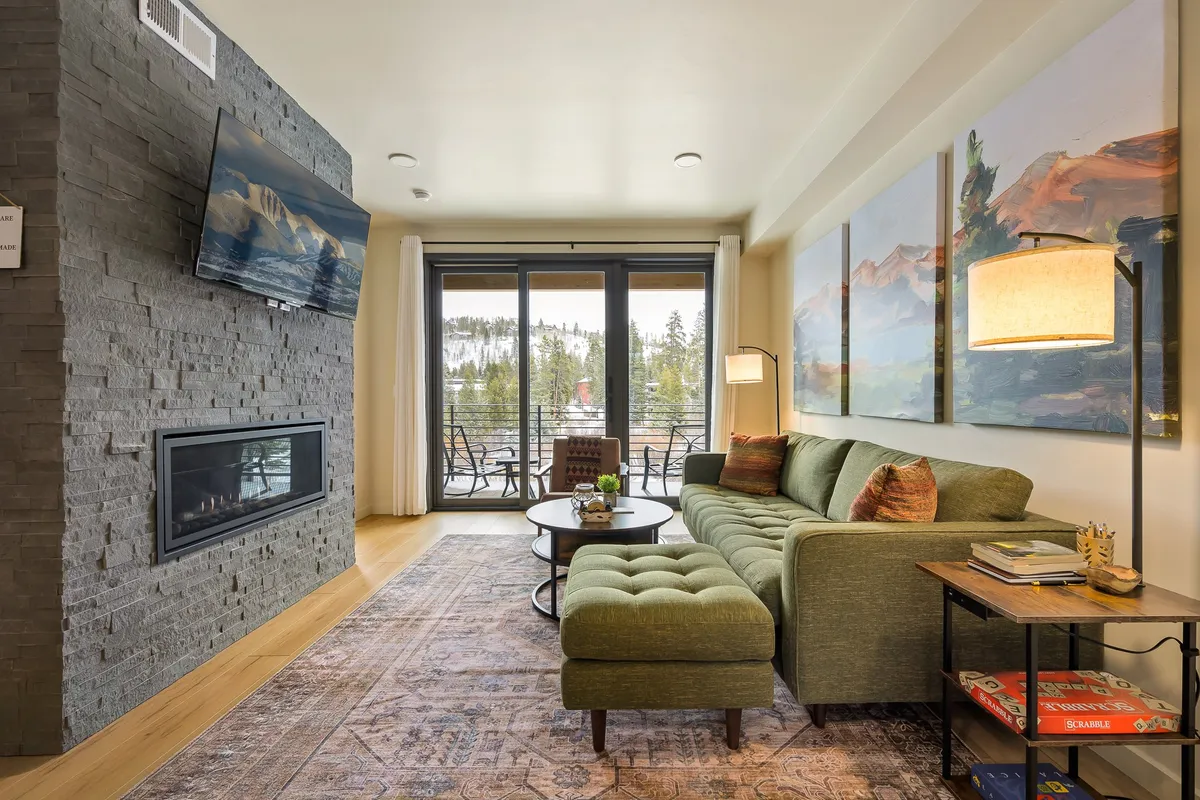 The living area is centered around a stylish green sectional sofa and a cozy fireplace, with large windows offering scenic outdoor views. Abstract art and natural light create a warm, comfortable space ideal for family relaxation, reading, or gathering together.